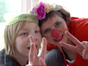 Ein Clown mit roter Nase und ein Kind zeigen das Peace-Zeichen in die Kamera.