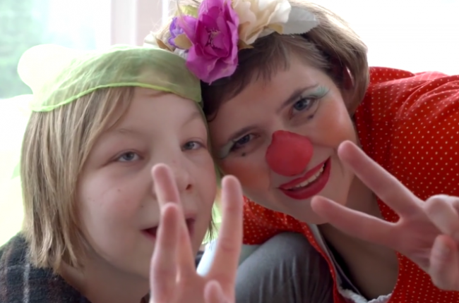 Ein Clown mit roter Nase und ein Kind zeigen das Peace-Zeichen in die Kamera.