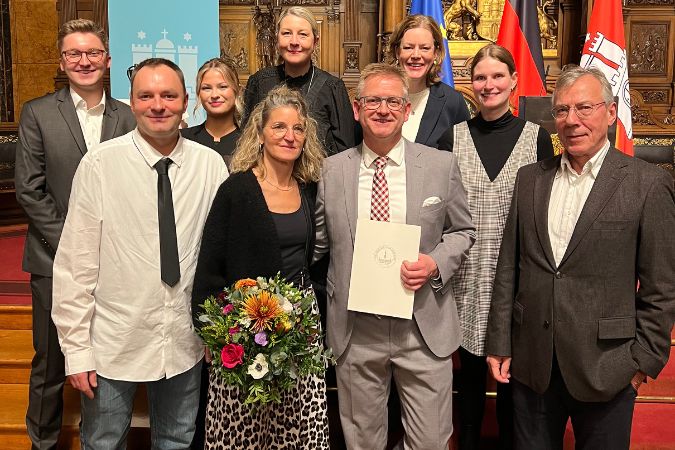 Eine Gruppe von Personen posiert für ein Foto bei einer Preisverleihung mit einer Urkunde und einem Blumenstrauß.