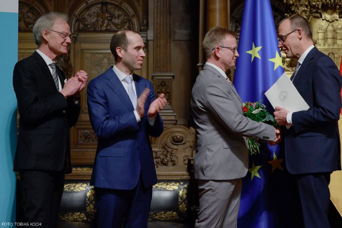 Friedrich Merz überreicht eine Urkunde und Blumen an Steffen Schumann. Zwei weitere Männer stehen daneben und klatschen.