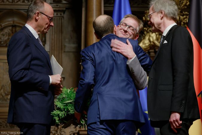 Vier Männer stehen auf einer Bühne, einer umarmt einen anderen und hält einen Blumenstrauß.