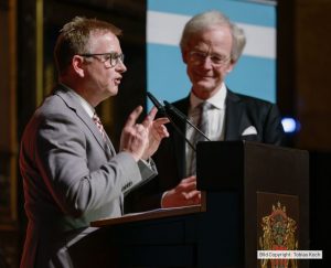 Zwei Männer stehen an einem Rednerpult während einer Preisverleihung, einer spricht, der andere hört zu.