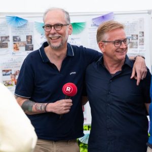 Zwei Männer stehen nebeneinander und lächeln, einer hält ein Mikrofon in der Hand.