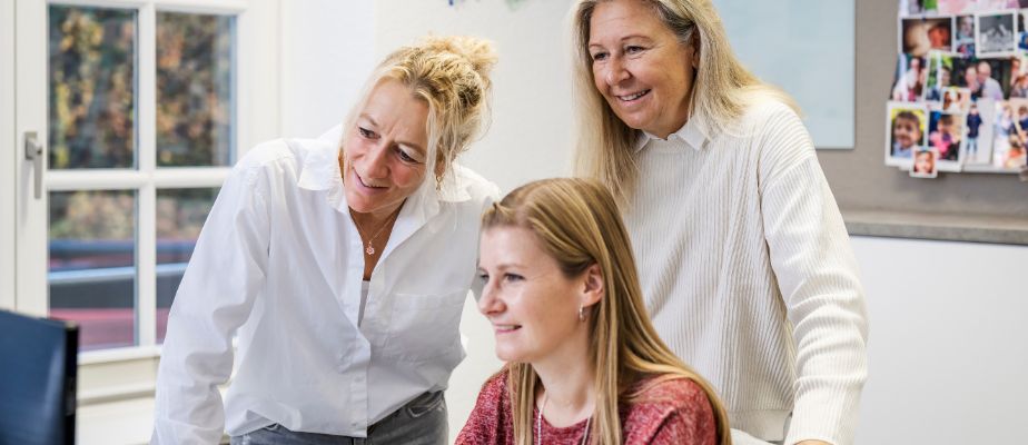 Drei Frauen, die Kolleginnen aus dem Familienbüro, stehen zusammen an einem Schreibtisch und gucken auf einen Bildschirm.