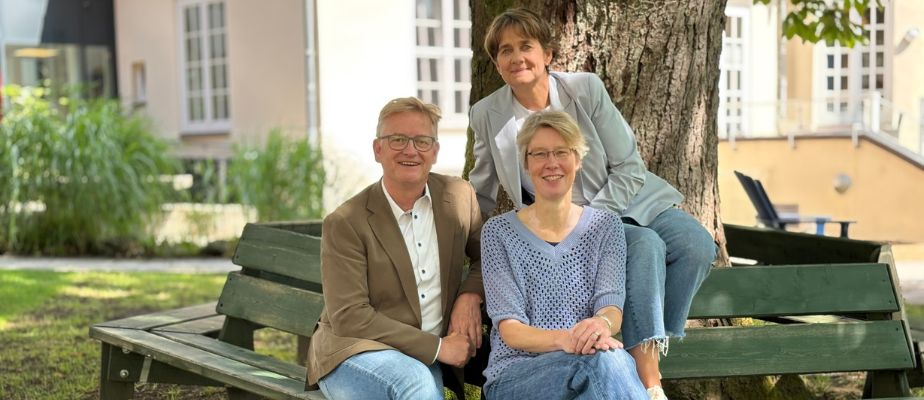 Steffen Schumann, Andrea Jaap und Ute Wöllmann sitzen auf einer grünen Bank vor einem Baum. Alle drei lächeln in die Kamera.