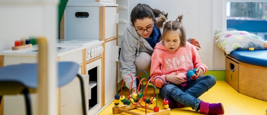 Eine junge Pflegekraft sitzt mit einem Gastkind in unserem Aufenthaltsbereich der Kinderetage. Sie spielen zusammen an einer Motorikschleife.