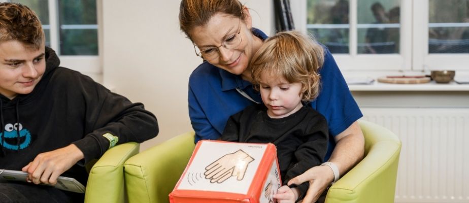 Eine Pflegekraft sitzt auf einem grünen Sessel und hat ein Gastkind auf dem Schoß. Der kleine Junge hat einen sehr großen Würfel mit Symbolen und Bildern auf dem Schoß. Beide schauen den Würfel an. Neben den beiden sitzt ein weiterer, etwas älterer Junge und schaut den beiden zu.