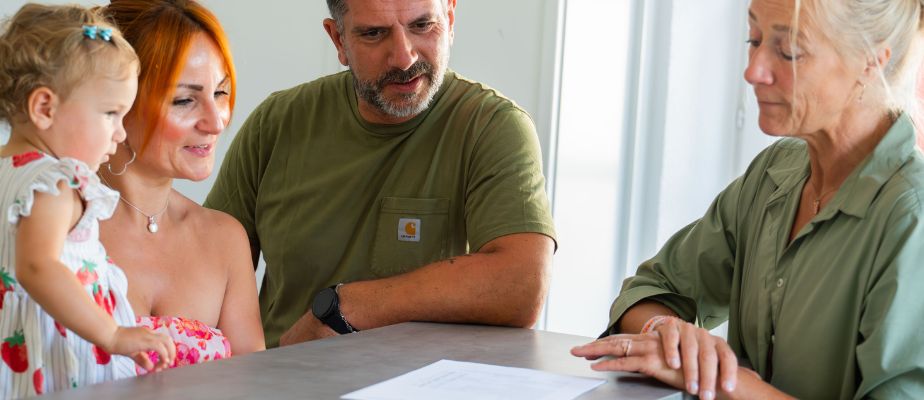 Eine Familie, bestehend aus einem Mann, einer Frau und einem kleinen Mädchen, steht in unserem Familienbüro an einem Tisch. Neben ihnen steht eine Kollegin aus dem Familienbüro. Alle schauen auf einen Zettel, der auf dem Tisch liegt.