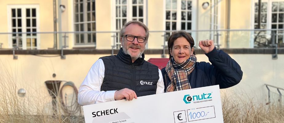 Ein Mann und eine Frau stehen vor einem Gebäude und halten einen Scheck mit einer 1000 € Spende für Kinder mit Behinderung in der Hand.