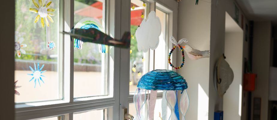 Der Blick durch das Fenster vom Kunstraum in den Garten des Neuen Kupferhofs. Das Fenster ist mit Window Color bemalt und vor dem Fenster hängen ein gebasteltes Flugzeug, eine Wolke, eine Qualle, ein Armband und ein Vogel.