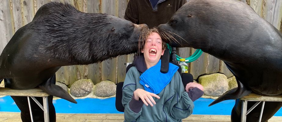 Ein Mädchen im Rollstuhl sitzt zwischen zwei Seelöwen. Die beiden Seelöwen geben dem Mädchen einen Kuss auf den Kopf. Das Mädchen hat den Mund freudig aufgerissen.