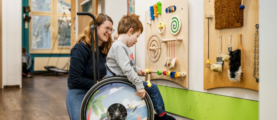 Ein kleiner Junge im Rollstuhl sitzt vor einer Spieltafel auf der Kinderetage. Links neben ihm kniet seine Mutter und lächelt.
