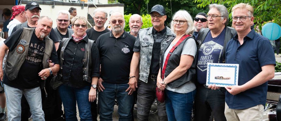 Steffen Schumann steht neben einer Gruppe von elf Harleyfahrer:innen und hält einen Spendencheck in der Hand.