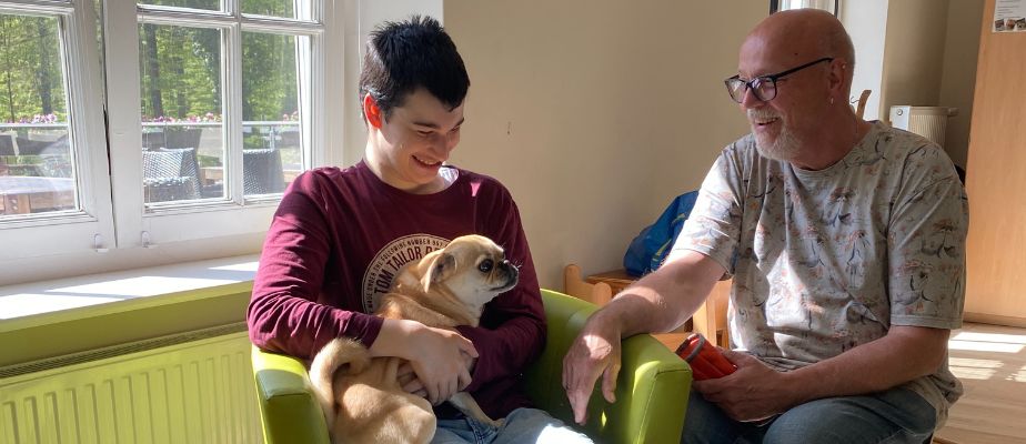 Ein Junge sitzt auf einem grünen Sessel und hat einen Mops auf dem Schoß. Neben ihm sitzt ein Mann, der die beiden anguckt. Beide lachen.