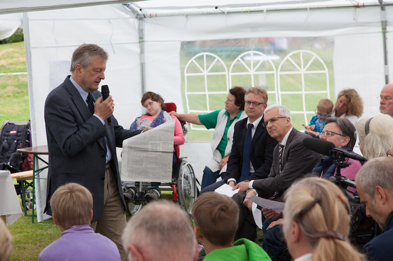 Ein Mann hält eine Rede in einem Zelt, während das Publikum aufmerksam zuhört.