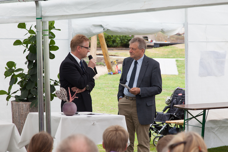 Zwei Männer stehen in einem Zelt und führen ein Gespräch, während eine Pflanze im Hintergrund sichtbar ist.