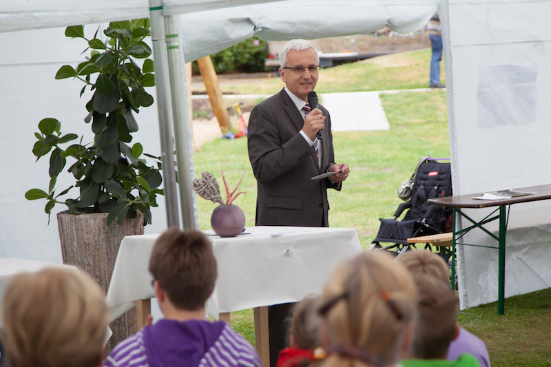 Ein Mann spricht mit einem Mikrofon während einer Veranstaltung im Freien, mit Blick auf das Publikum.