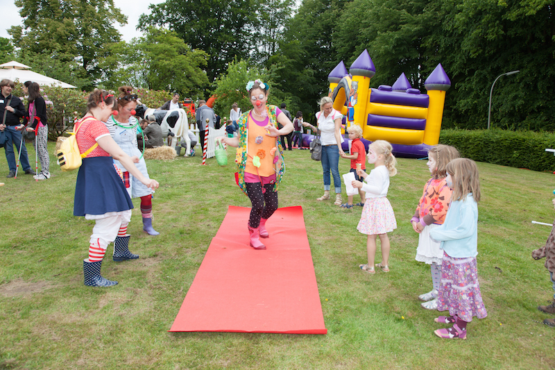 Clown in buntem Kostüm, der auf einem roten Teppich bei einer Veranstaltung läuft, umgeben von Kindern und anderen Personen.