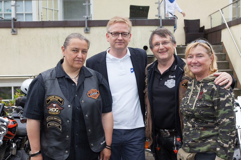 Vier Personen stehen zusammen, während Motorräder im Hintergrund sichtbar sind.