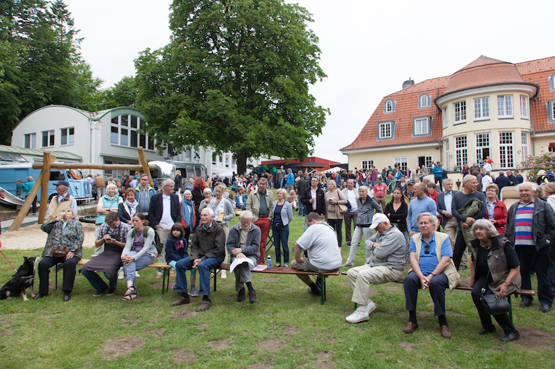 Eine große Menschenmenge sitzt und steht bei einer Veranstaltung im Freien, umgeben von Bäumen und Gebäuden.