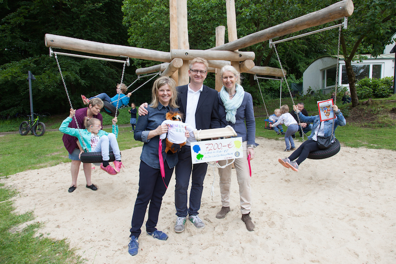 Drei Personen halten einen Scheck auf einem Spielplatz mit einer Holzspielstruktur im Hintergrund.