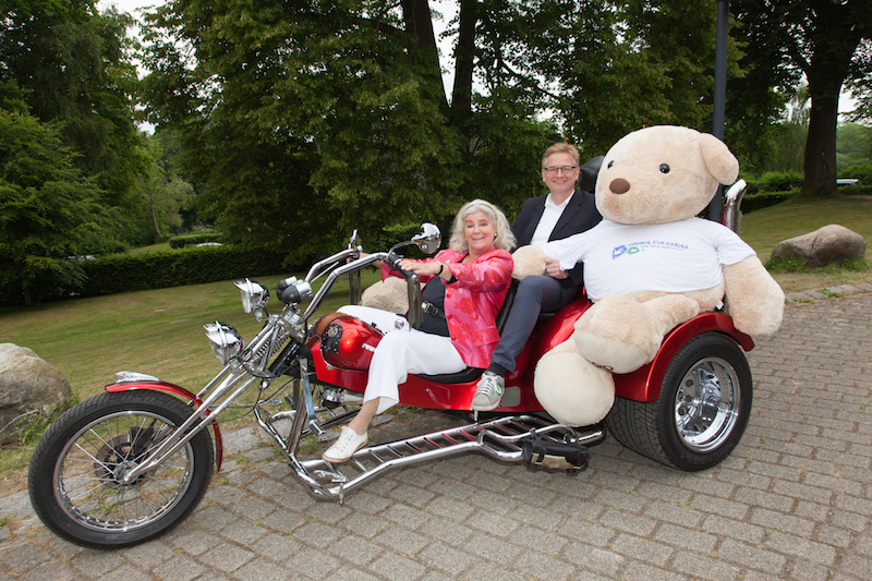Eine Frau und ein Mann sitzen auf einem roten Motor-Dreirad mit einem großen Teddybären auf dem Beifahrersitz.