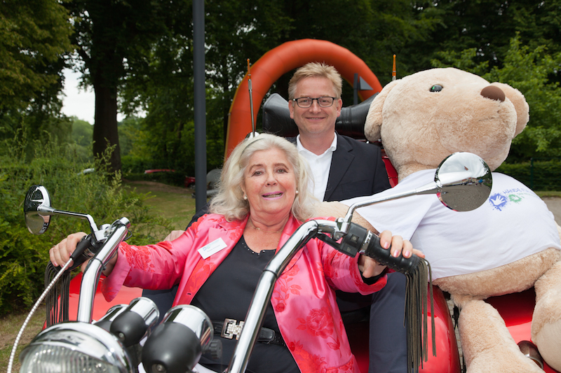 Frau in einem roten Blazer und Mann in Anzug sitzen auf einem Motorrad mit einem großen Bären im Hintergrund.