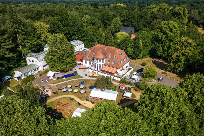 Luftaufnahme eines großen Gebäudes mit roten Dächern und umgebenden Gartenflächen.