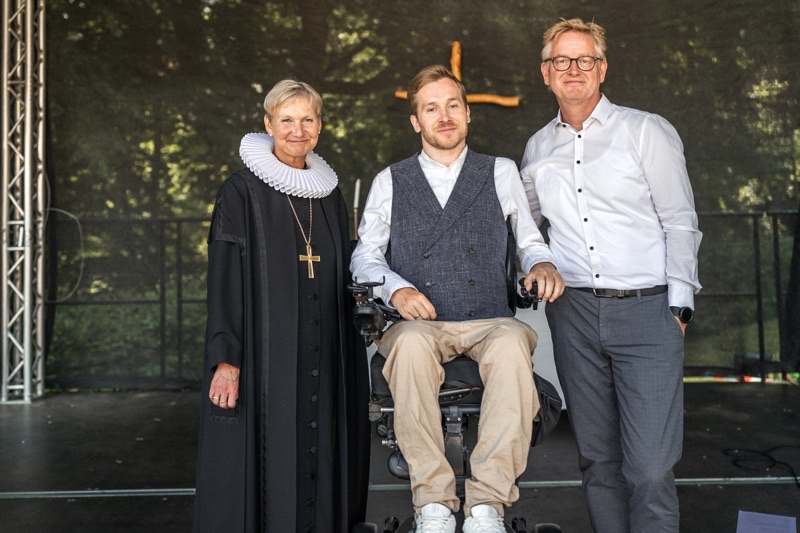 Drei Personen stehen auf einer Bühne, eine Frau in schwarzer Robe, ein Mann im Rollstuhl und ein Mann in einem weißen Hemd.