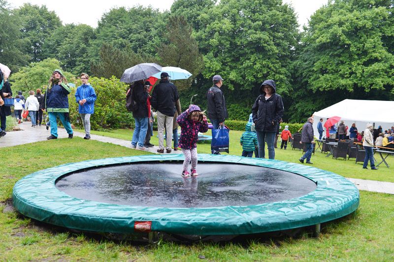 Ein Kind steht auf einem nassen Trampolin im Freien. Im Hintergrund viele Menschen.