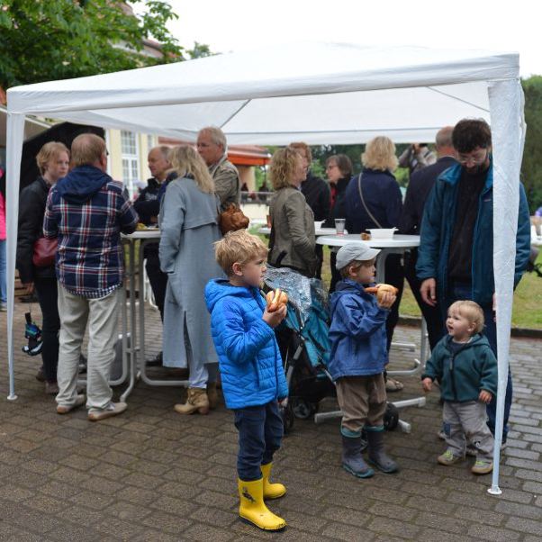 Drei Kinder stehen unter einem Pavillon und essen umgeben von Erwachsenen.
