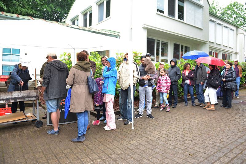 Eine Gruppe von Menschen steht in einer Schlange bei einem Essensstand, einige halten Regenschirme.