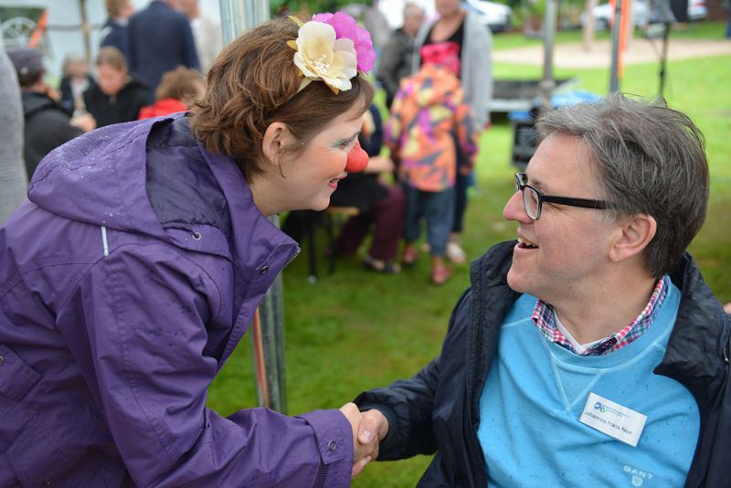 Eine Frau mit einer Clownsnase und Blumen im Haar schüttelt die Hand eines Mannes in einem blauen Pullover.