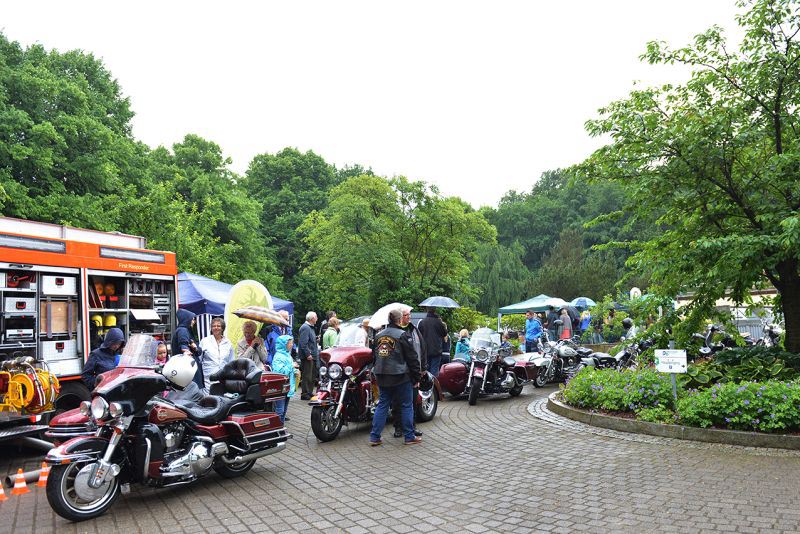 Reihe von Motorrädern auf einem Platz während eines regnerischen Tages mit Menschen, einem Feuerwehrauto und Ständen im Hintergrund.
