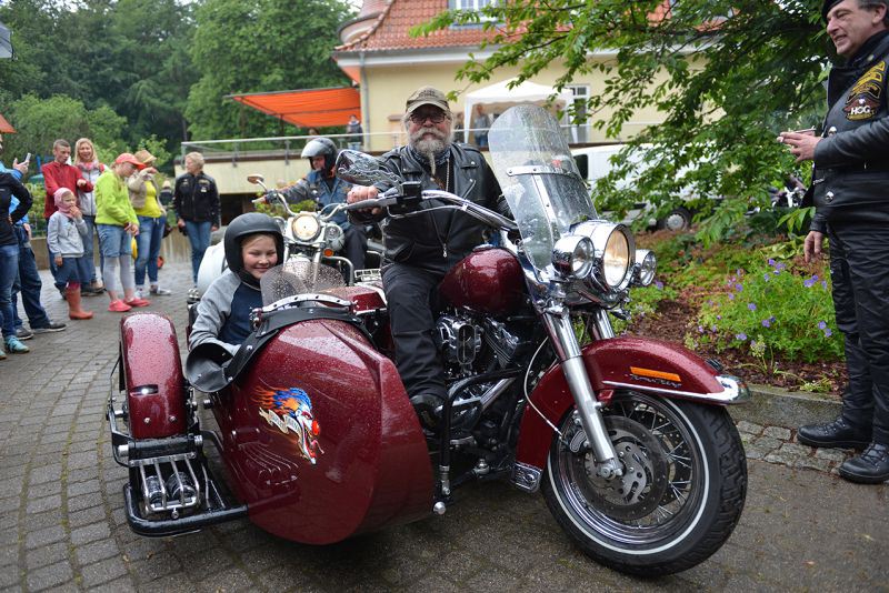 Ein Mann mit Bart sitzt auf einem roten Motorrad mit Beiwagen, während ein Kind neben ihm sitzt.