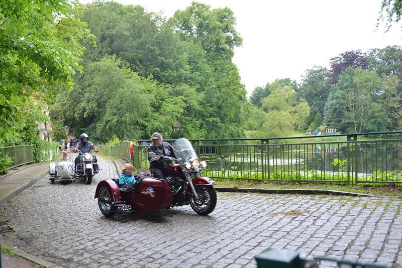 Ein rotes Motorrad mit Beiwagen fährt auf einer gepflasterten Straße entlang einer grünen Umgebung.