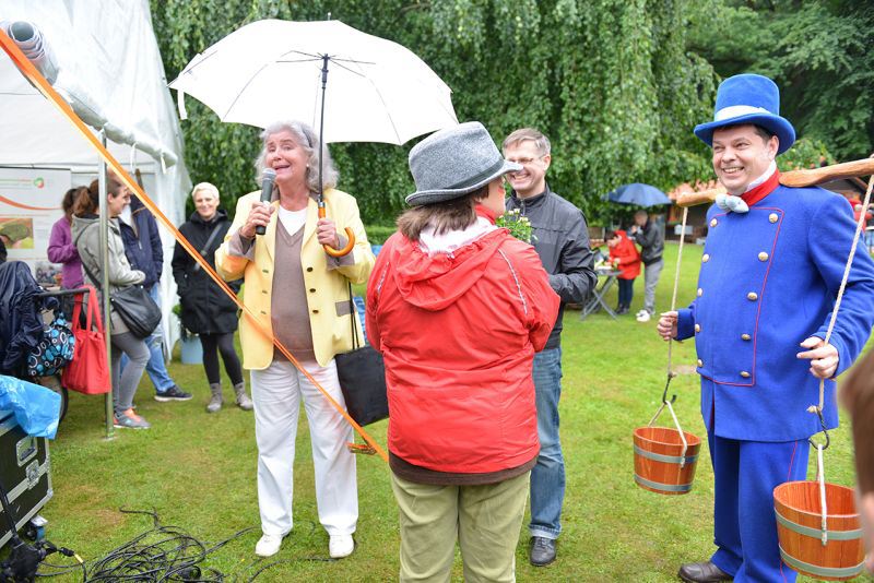 Eine Person mit einem Regenschirm spricht zu einer Gruppe, während ein Mann in einem blauen Kostüm mit Eimern daneben steht.