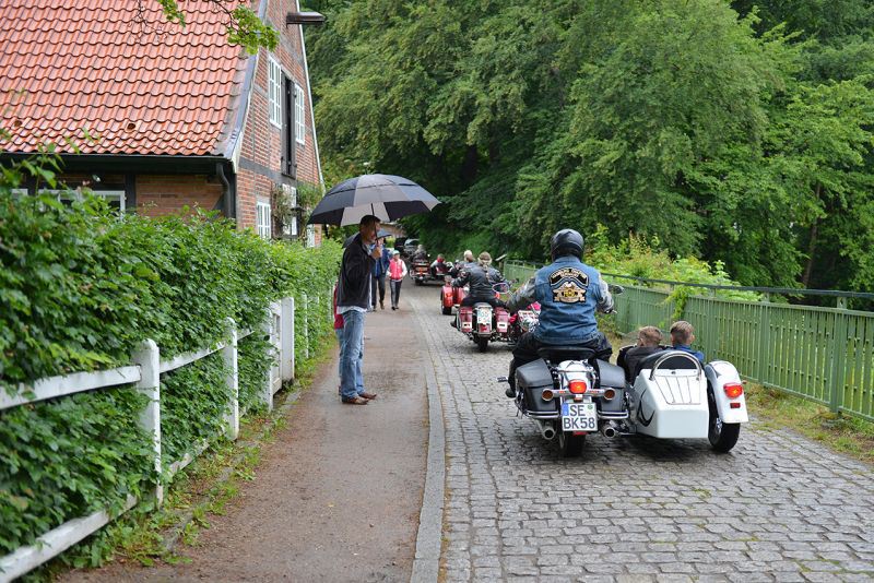 Gruppe von Motorradfahrer:innen mit Beiwagen, in denen weitere Personen sitzen auf einer gepflasterten Straße, während eine Person mit einem Regenschirm auf dem Gehweg steht.