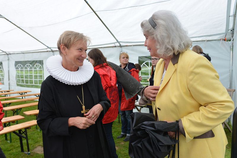 Zwei Frauen, eine in schwarzer Robe mit einem weißen Kragen und die andere in einem gelben Jackett, unter einem Zelt im Gespräch.
