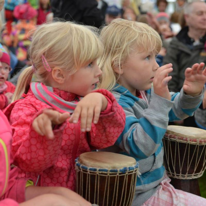Zwei blonde Kinder mit Trommeln, die aufmerksam auf eine Darbietung schauen.