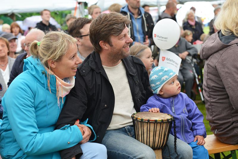 Eine Familie mit einem Kind sitzt auf einer Bank. Vor ihnen steht eine Trommel.
