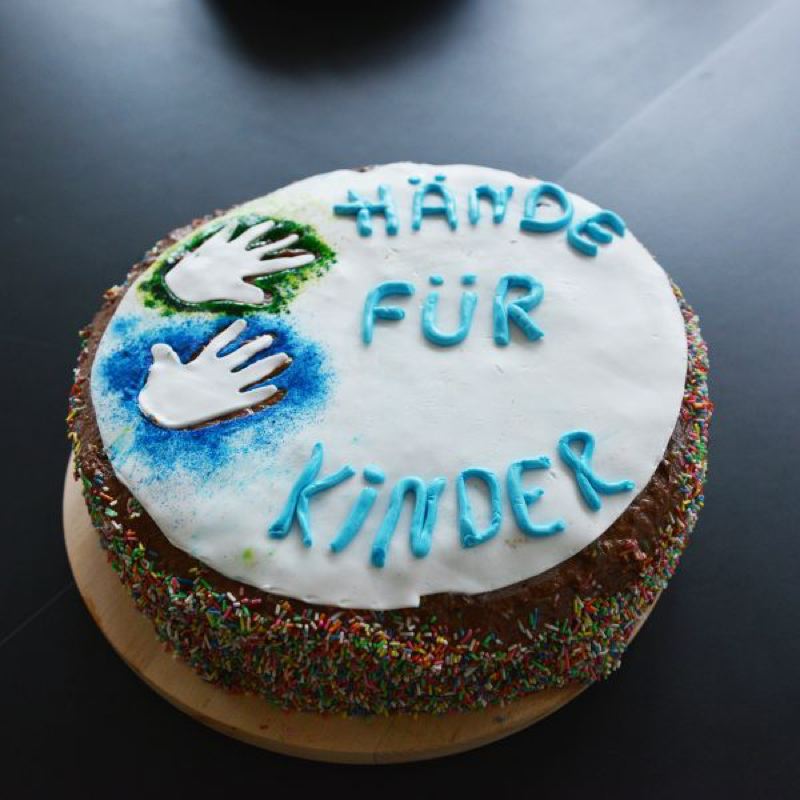 Runder Kuchen mit weißem Zuckerguss, blauen und grünen Handabdrücken sowie bunten Streuseln.
