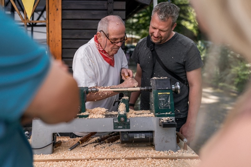 Zwei Männer arbeiten gemeinsam an einer Holz-Drehmaschine, während Späne um sie herum liegen.