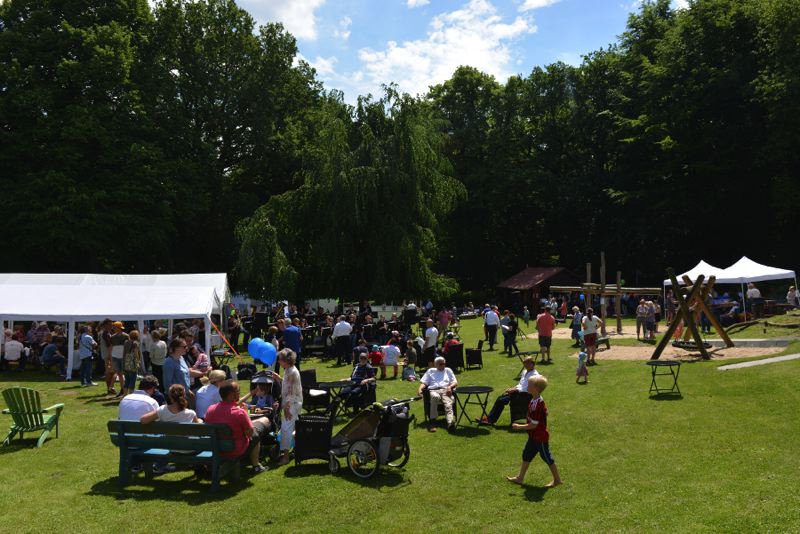 Menschenmenge auf einem Sommerfest im Garten mit Zelten und Spielplatz im Hintergrund.