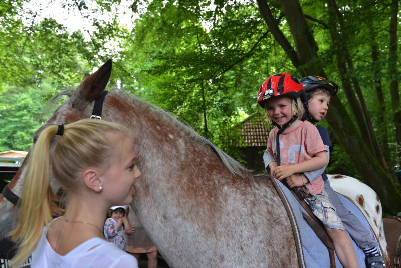 Zwei Kinder mit Helmen sitzen auf einem Pferd, während eine Person daneben steht.