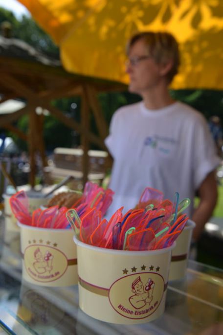 Zwei Eis-Becher mit bunten Löffeln, im Hintergrund eine Person im T-Shirt.