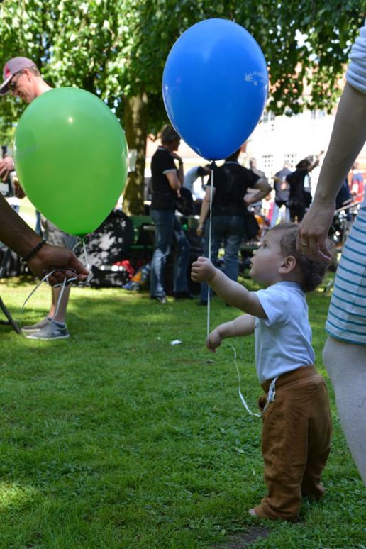 Ein kleines Kind steht auf Rasen und hält einen blauen Luftballon in der Hand. Auch ein grüner Ballon ist zu sehen.