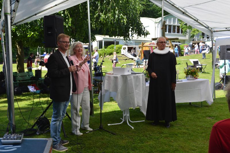Drei Personen stehen unter einem Zelt während einer Veranstaltung im Freien, zwei sprechen ins Mikrofon.