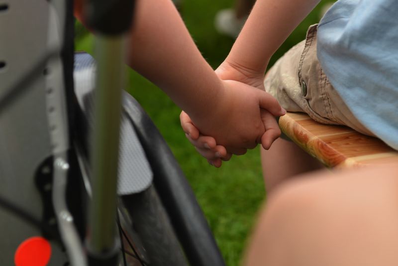 Zwei Kinderhände, die sich festhalten, ein Kind sitzt im Rollstuhl, das andere auf einer Bank.