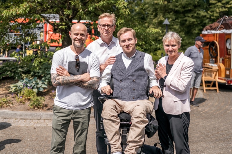 Vier Personen posieren gemeinsam für ein Foto im Freien, eine Person im Rollstuhl.